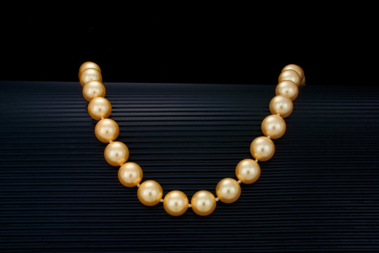 A Single Strand Golden Color Pearl Necklace Drapes Over A Curve Black Ribbed Background.