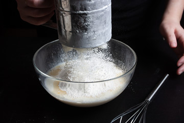 The cook sprinkles flour through a sieve into a glass bowl with milk and eggs to prepare a delicious meal. Pancake Recipe. Dark background.