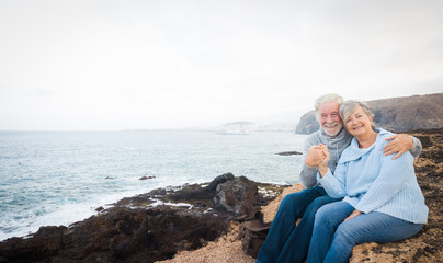 Fototapeta na wymiar Couple of senior people sitting on the cliff of the ocean and looking at camera Hugging and smiling. Morning soon outside, ready for un healthy excursion. Vacation and happiness