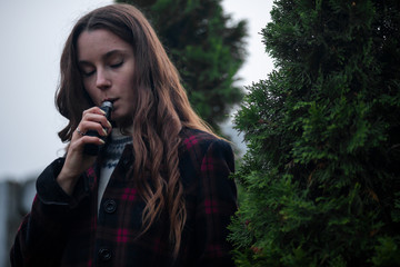 Vape teenager. Young pretty white girl in checkered coat smoking an electronic cigarette near the thuja on the street in the park in the autumn evening. Bad habit. Vaping activity.