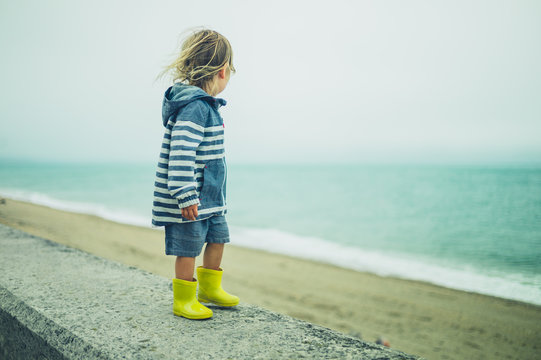 Little toddler in weathreproofs standing on wall by sea