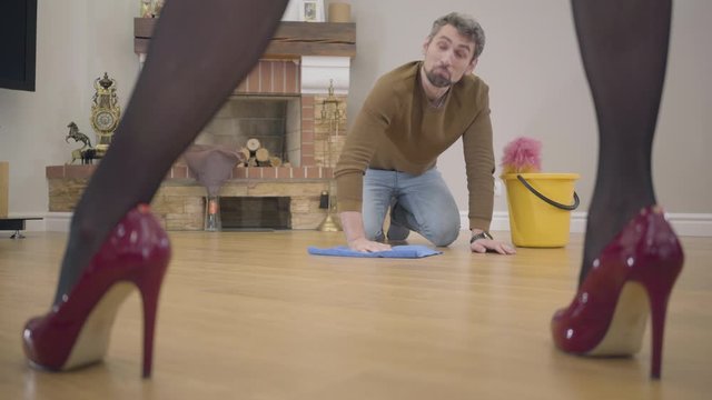 Portrait of adult Caucasian guy washing the floor and talking to his wife or girlfriend. Beautiful female legs in elegant red high-heel shoes on the foreground. Guilty henpecked doing housework.