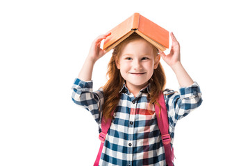 cheerful redhead pupil holding orange book above head isolated on white
