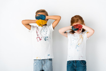 cute kids with paint on hands covering faces while standing on white