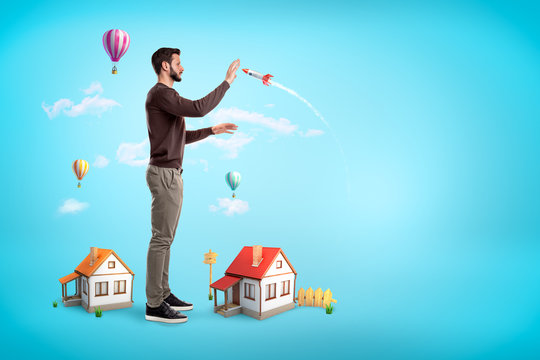 Side view of giant young man trying to catch space rocket to protect two small houses at his feet, on blue background with copy space.
