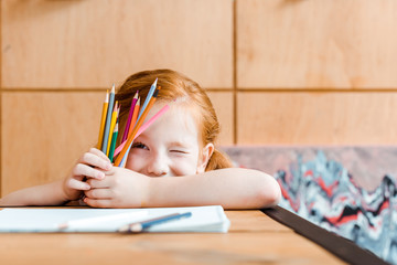 selective focus of cute redhead kid winking eye while holding color pencils