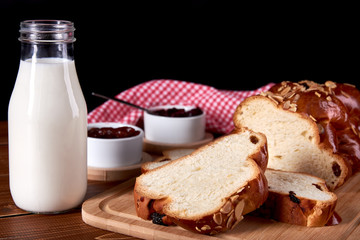 Still life on old wooden table top. Knitted Christmas cake or sweet pastry with raisins and almond slices. Bowl of fruit jam and bottle of milk on black background.