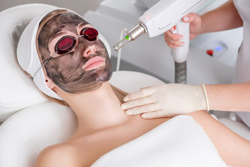 Naklejka premium Close-up of a cosmetologist's hand making a hardware carbon peeling by a young pretty customer woman in safety glasses and a headdress lying on a couch in a beauty salon. Facial cleansing concept