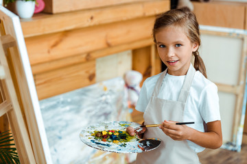 happy kid in apron holding palette with colorful gouache paint