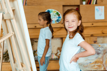 selective focus of happy redhead kid standing with hand on hip near child in art school