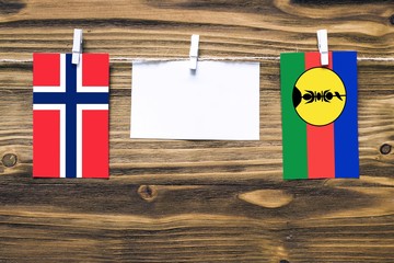 Hanging flags of Bouvet Islands and New Caledonia attached to rope with clothes pins with copy space on white note paper on wooden background.Diplomatic relations between countries.