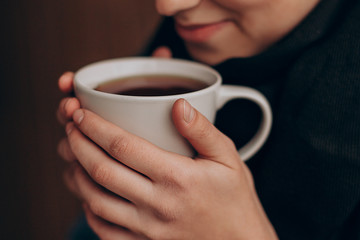 a mug of hot coffee or tea in the hands of a young pretty girl near her lips. Cropped photo with focus on cup