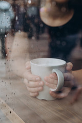 Woman hand holding the cup of coffee or tea on rainy day window background in vintage color tone, copy space