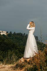 Beautiful elegant bride in lace wedding dress with long full skirt and long sleeves. Pretty girl in white. Nature, with city in the background.