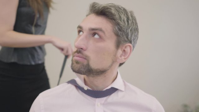 Close-up portrait of scared young Caucasian henpecked man. Wife or girlfriend in leather clothes walking at the background with swatter. Husband or boyfriend controlled by his spouse.