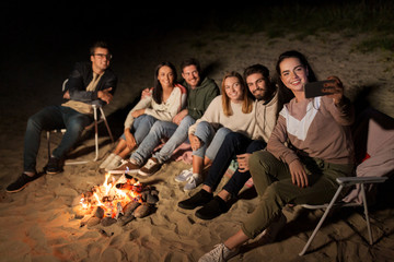 leisure and people concept - group of smiling friends sitting at camp fire taking selfie by smartphone on beach at night