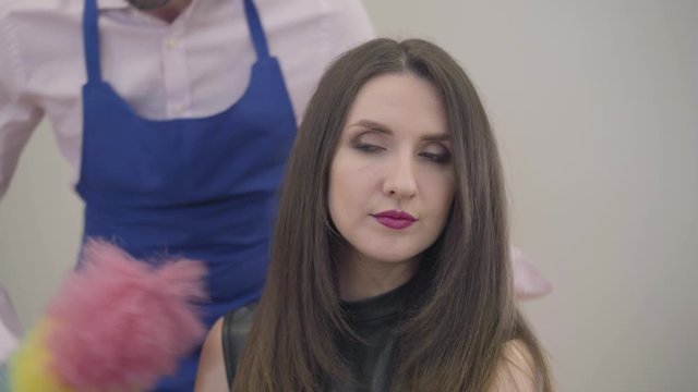 Portrait of a young beautiful Caucasian woman with long brunette hair and brown eyes. Man with dust whisk coming up and putting crown on her head. Strong wife controlling henpecked husband.