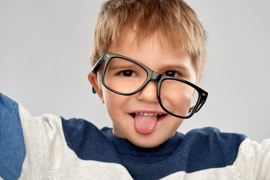 school, education and vision concept - portrait of smiling little boy with crookedly worn glasses showing tongue over grey background