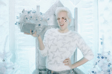 christmas unhappy sad blonde caucasian guy in white sweater with stars holding and opening gift present on the chair