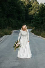 Beautiful elegant bride in lace wedding dress with long full skirt and long sleeves. She is holding a big bouquet of flowers. Outdoors, on the road.
