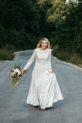 Beautiful elegant bride in lace wedding dress with long full skirt and long sleeves. She is holding a big bouquet of flowers. Outdoors, on the road.