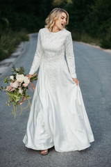 Beautiful elegant bride in lace wedding dress with long full skirt and long sleeves. She is holding a big bouquet of flowers. Outdoors, on the road.