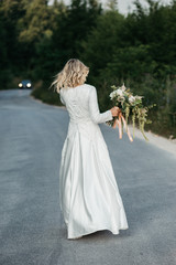 Fototapeta na wymiar Beautiful elegant bride in lace wedding dress with long full skirt and long sleeves. She is holding a big bouquet of flowers. Outdoors, on the road.