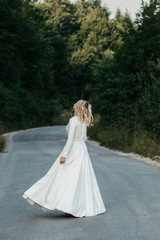 Fototapeta na wymiar Beautiful elegant bride in lace wedding dress with long full skirt and long sleeves. She is holding a big bouquet of flowers. Outdoors, on the road.