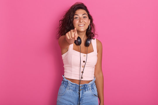 Close up portrait of pretty girl isolated over pink studio background, poiting with her index finger to camera, charming female wearing rose top and jeans, hacing dark hair, girl expressing happyness.