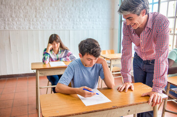 Confident caucasian teenager boy in the classroom doing school test and talking to his teacher