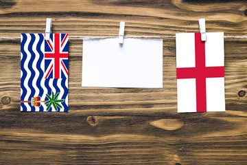 Hanging flags of British Indian Ocean Territory and England attached to rope with clothes pins with copy space on white note paper on wooden background.