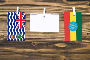 Hanging flags of British Indian Ocean Territory and Ethiopia attached to rope with clothes pins with copy space on white note paper on wooden background.