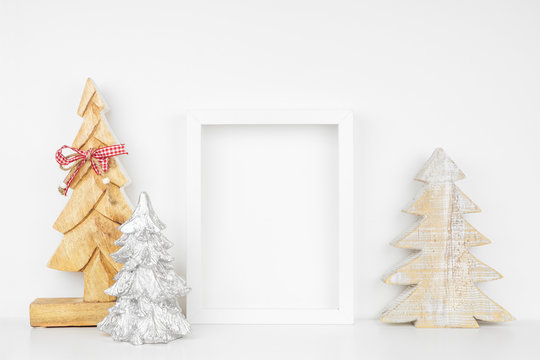 Mock up white frame with wooden Christmas tree decorations on a shelf. Portrait frame against a white wall.