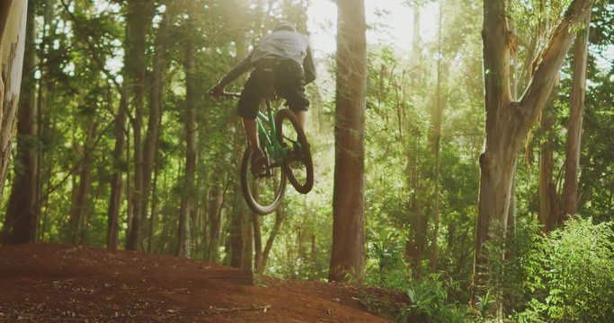 Young active mountain biker catching big air off of a wooden jump at sunset with a green forest background, epic mountain biking jump in slow motion