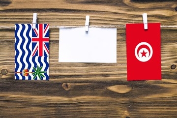 Hanging flags of British Indian Ocean Territory and Tunisia attached to rope with clothes pins with copy space on white note paper on wooden background.