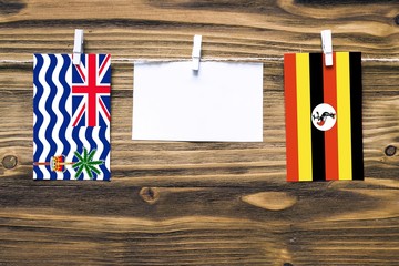 Hanging flags of British Indian Ocean Territory and Uganda attached to rope with clothes pins with copy space on white note paper on wooden background.