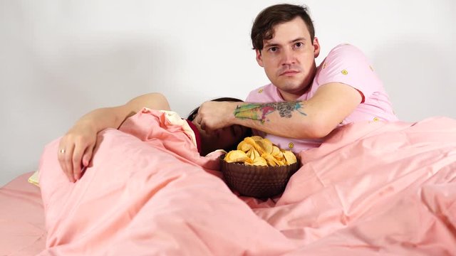 A young married couple are lying in bed, watching a film and eating chips. The husband feeds his wife chips. The woman is falling asleep from the boring movie.