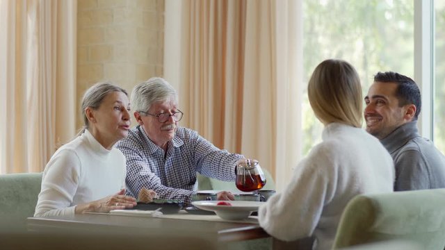 Tracking medium shot of young and senior couples, two generation of family having dinner together in restaurant. Elderly man pouring tea from pot for his wife and son with girlfriend
