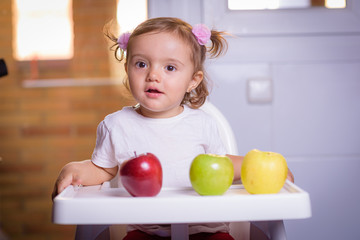 Cute baby 1,4 years old sitting on high children chair and eating fruit alone in white and brown kitchen