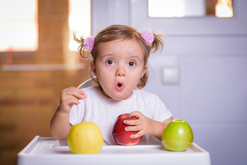 Fototapeta premium Cute baby 1,4 years old sitting on high children chair and eating fruit alone in white and brown kitchen