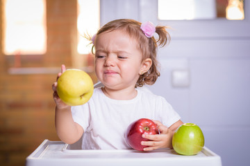 Fototapeta na wymiar Cute 1.4 year old baby sitting in a high chair for children angry because he does not like the apple.