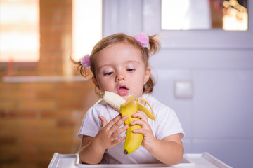 Cute baby 1,4 years old sitting on high children chair and eating a banana alone in white kitchen