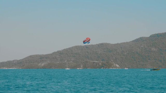 Boats on sea waves and person parasailing above water. Scenic view of beautiful sea harbour with vessels and person gliding through air wearing open parachute above motorboat