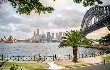 Breite Aufnahme von Touristen, die an einem Sommerabend entlang des Hafens von Sydney radeln, mit Blick auf die Skyline von Sydney, Milsons Point, Waterfront, Sydney Harbour Bridge und das Opernhaus - Sydney, Australien