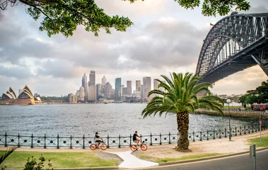 Photo sur Plexiglas Sydney Harbour Bridge Plan large de touristes faisant du vélo le long du port de Sydney, avec vue sur la ligne d& 39 horizon de Sydney, Milsons Point, le front de mer, le pont du port de Sydney et l& 39 opéra un soir d& 39 été - Sydney, Australie