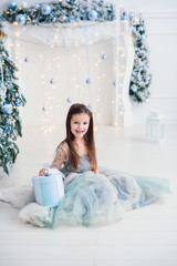 Cute little girl with gift box in beautiful dress sits near Christmas tree on fireplace backdrop.