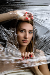 Abstract mysterious portrait girl hidden white behind breaking stretch cellophane and looking through the gap plastic. Pretty brunette wrapped in cellophane.