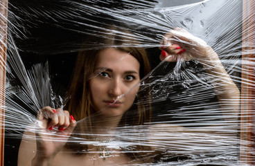 black and white Abstract mysterious portrait girl hidden white behind breaking stretch cellophane and looking through the gap plastic. Pretty brunette wrapped in cellophane.