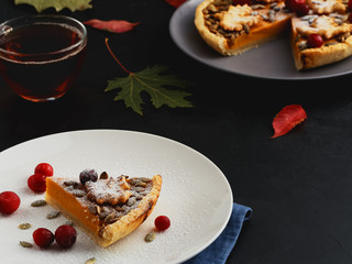 A piece of homemade traditional American pumpkin pie is located on a plate. Close-up. On a black background multi-colored autumn leaves and a cup with tea. Selective focus.