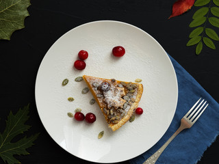 A slice of homemade traditional American pumpkin pie decorated with icing sugar and cranberries is located on a white plate. Top view. On a black background colorful autumn leaves.